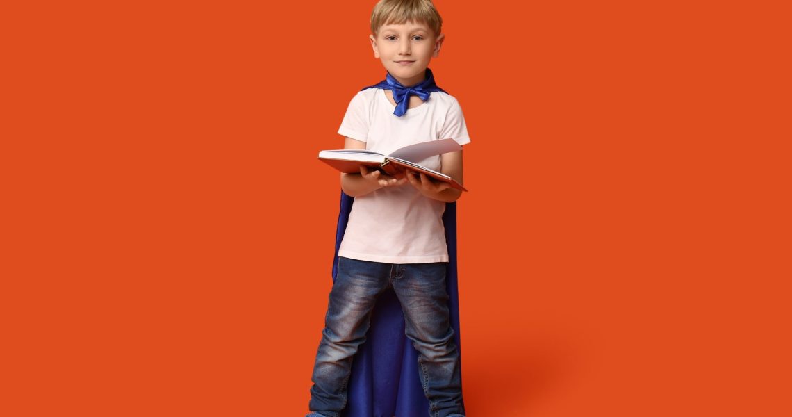 Cute,Little,Boy,Dressed,As,Superhero,With,Book,On,Orange